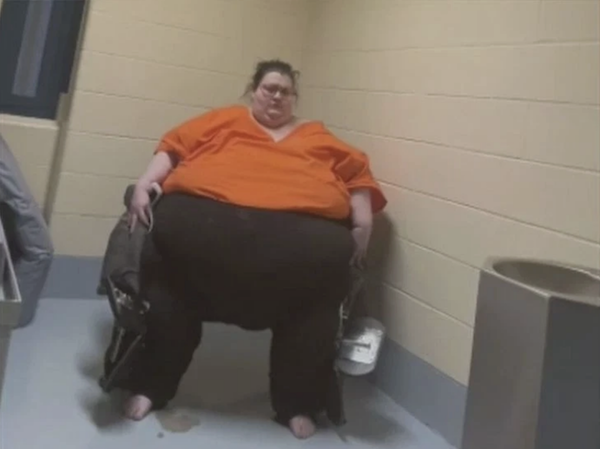 An individual in an orange shirt and dark pants is seated in a wheelchair in a bare room, with a metal sink visible on the right and tiled walls surrounding them.