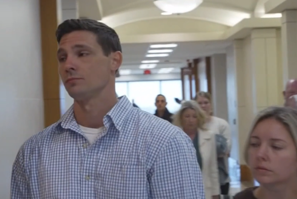 A solemn man leads a group of people through a courthouse hallway, light streaming in from windows in the background.