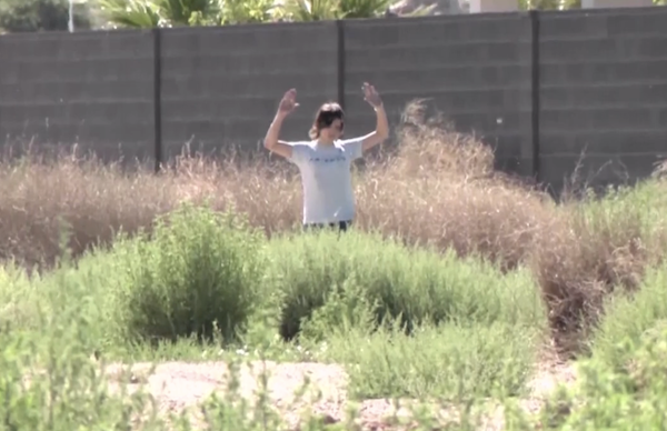 Someone in casual clothing raises their hands in a weedy vacant lot, positioned in front of a high, solid fence on a sunny day.