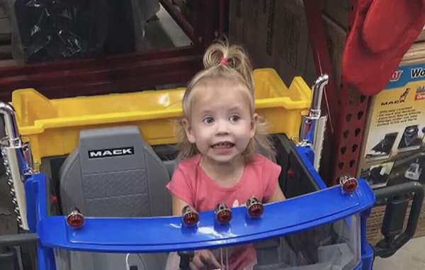 Child wearing a pink outfit poses cheerfully in a large toy truck cab, with store aisles visible in the background.
