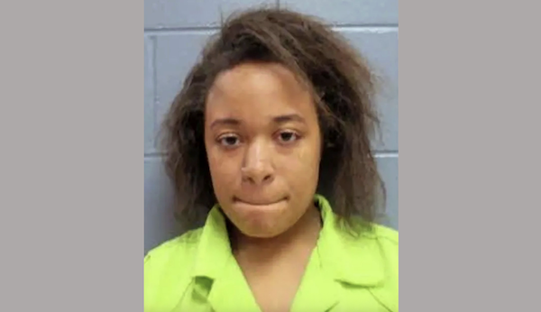 A woman with shoulder-length hair and a serious look on her face is shown in what appears to be a police booking photo, with a blue, block-patterned wall behind her.