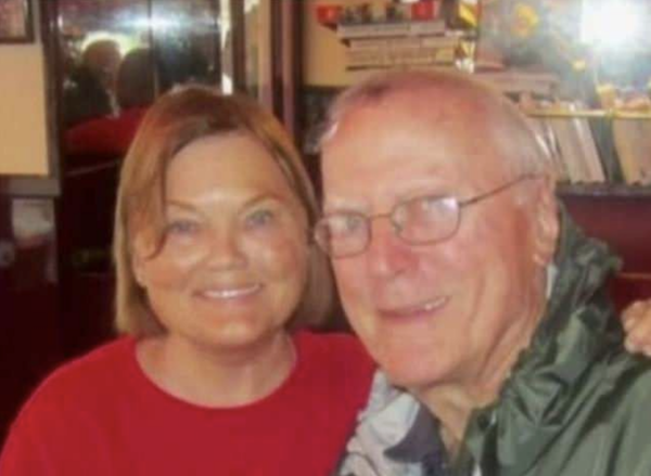 Cheerful middle-aged woman and older man, both seated and facing the camera, enjoy time together in a cozy indoor space with various items on shelves behind them.