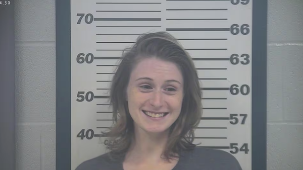 Female subject displaying a broad smile for a police booking photo, posed before a wall-lined height chart.
