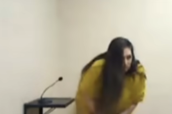 Woman dressed in yellow, possibly an inmate, holding papers while preparing to speak into a microphone in a minimalistic room with one chair.