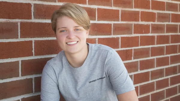 Person in a gray Under Armour shirt with short light hair sits against a brick wall, looking at the camera with a friendly expression.