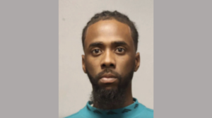 Front-facing photograph of a bearded man with dark hair, wearing a teal garment, with a neutral background.