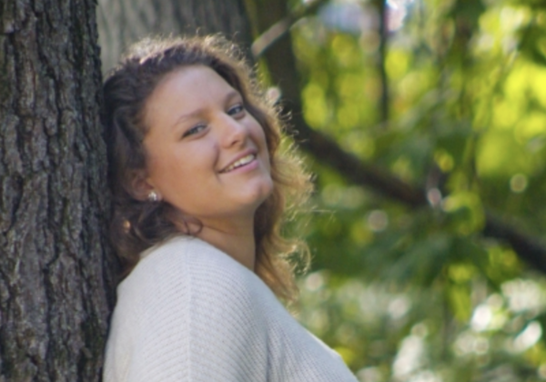 Side view of a smiling person in a pale sweater, posed against a tree with soft-focus leaves and branches in the background.