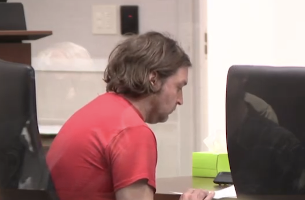 Person with slightly tousled hair wearing red attire sits at a wooden table, focusing downward, with blurred office or courtroom setting behind him.