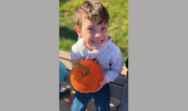 Happy child outdoors holding a pumpkin, dressed in a light-colored hoodie, with a blurred grassy background and sunlight highlighting his face.