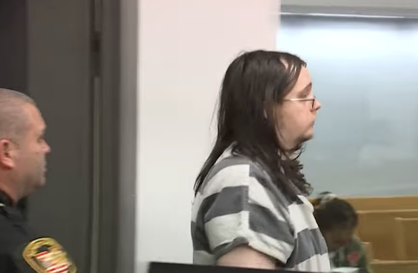 Side view of a person in jail clothing entering a courtroom, accompanied by a police officer.