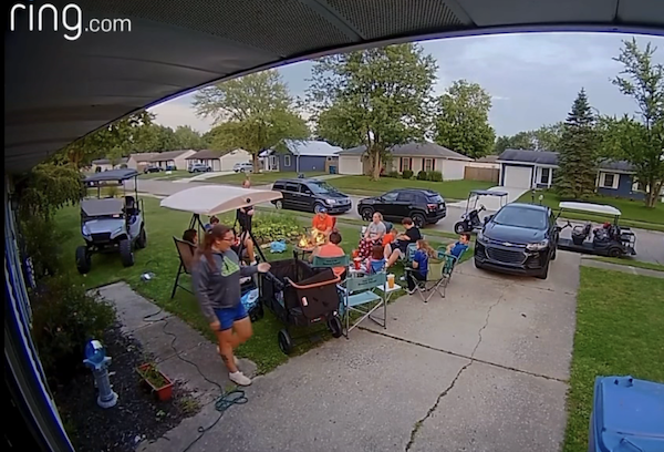 A casual neighborhood get-together shows people around a fire pit with drinks and snacks, parked cars and golf carts, and single-story homes visible across the street.