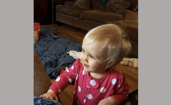 Small child with blonde hair and a cheerful expression wears a pink polka-dotted top, standing in a living space with a brown couch and some toys in the background.