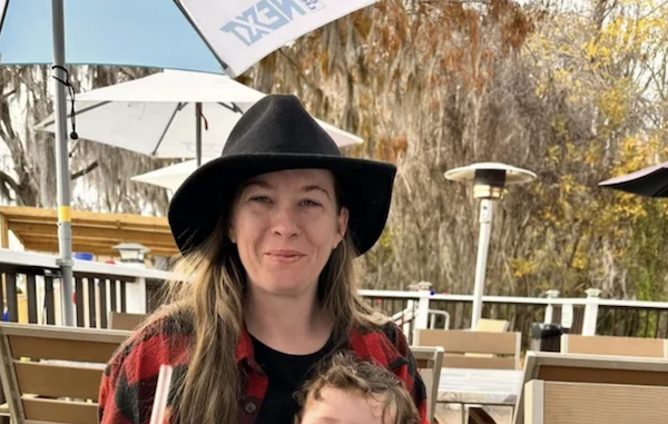 Outdoors at a patio restaurant, a woman dressed in a black felt hat and plaid shirt holds a curly-haired child, with parasols and outdoor seating behind them amidst autumn trees.