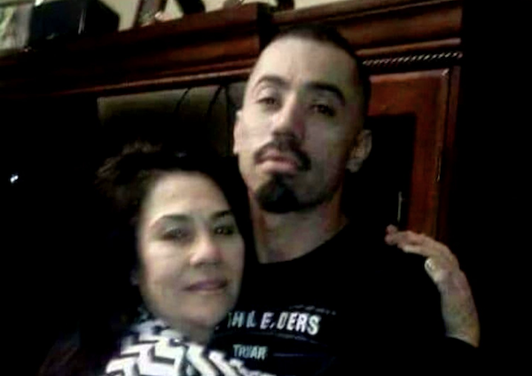 A man with facial hair and a woman with shoulder-length hair are pictured in an affectionate pose inside a home, with the background showing dark wooden cabinetry.
