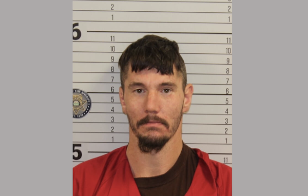 Head-and-shoulders image of a person with a trimmed beard and mustache, in front of a wall used for police mugshots, wearing a red shirt.