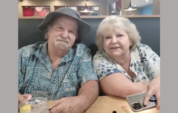 Smiling older man and woman are seated side by side in a restaurant, with drinks and a phone on the table in front of them.