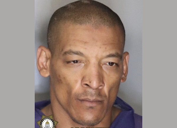 Front-facing photo of a person taken by police, with short hair, plain clothing, and sheriff’s insignia and “ARRESTED” text overlaying the corner.