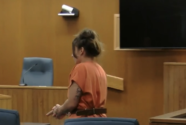 A woman with her hair in a bun, dressed in an orange outfit, stands alone in a courtroom, facing forward as empty seats surround her.