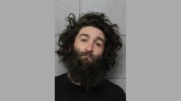 Arrest photo of a male individual with wild hair and prominent facial hair, looking slightly off-center before a tiled wall.