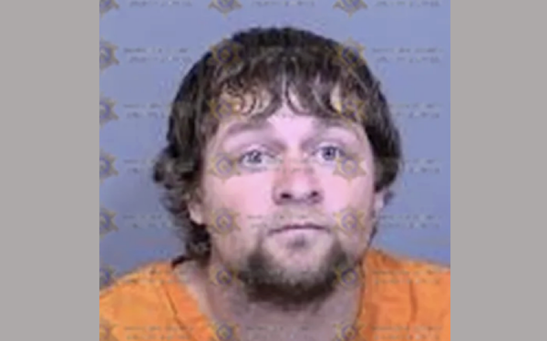 Portrait-style arrest photo of a man with a round face, blue eyes, and medium-length wavy hair, clad in an orange uniform, over a backdrop stamped with sheriff’s office logos.