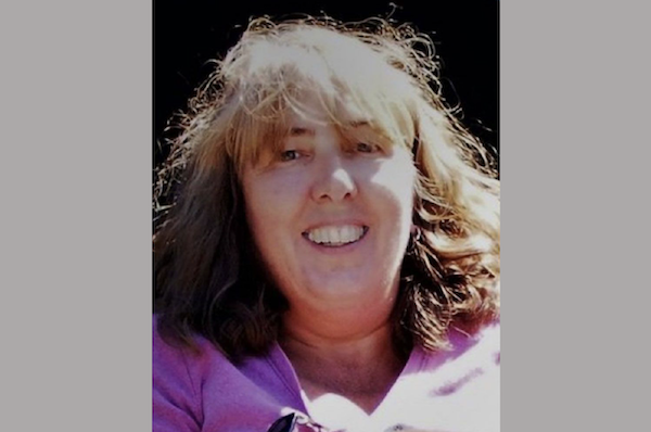A candid photo of a smiling woman with natural, light brown hair, taken on a sunny day with a simple dark backdrop.