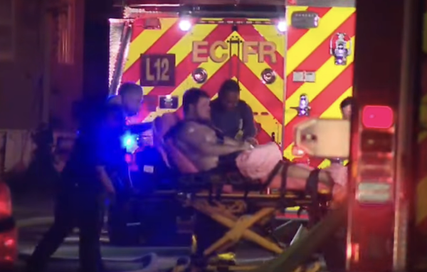 Rescue personnel assist a person lying on a stretcher, preparing to load him into a brightly lit Escambia County Fire Rescue ambulance outside a building at night.