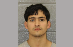 Identification photo showing a young adult male with dark hair and light skin, staring directly at the camera, background is gray.