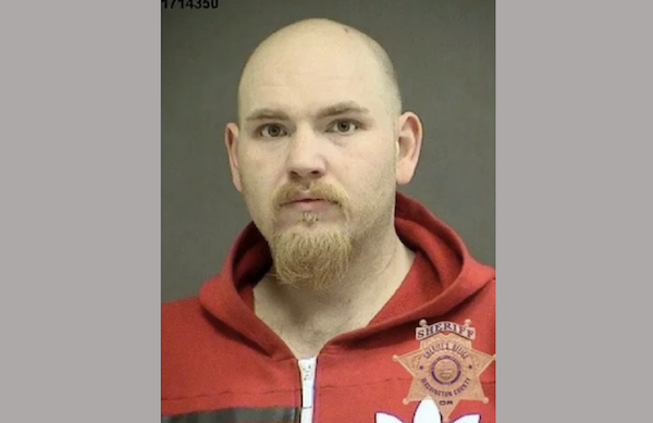 Portrait-style police photo showing a bearded individual in red outerwear, presented with a visible county sheriff's office insignia in the corner.