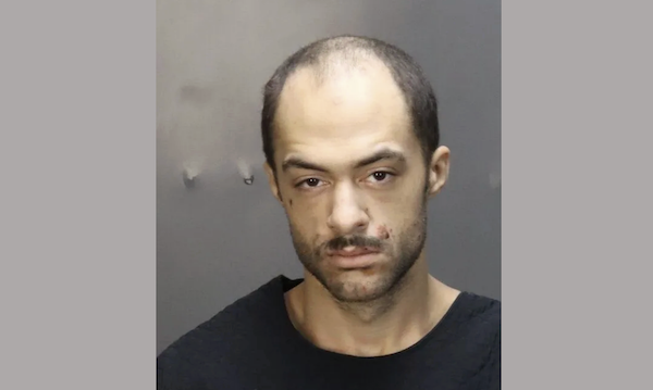 An image of a somber-looking man in a black shirt, photographed for a police mugshot against a gray background.