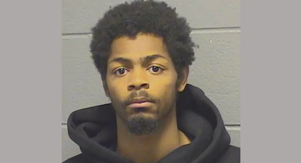 Young adult male with short afro and a goatee seen in a jail intake photograph, dressed in a black hoodie against a light gray brick wall.