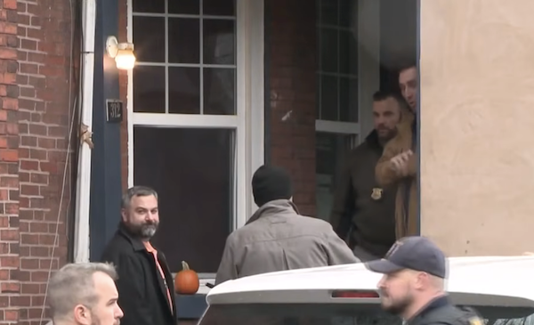 On a cloudy day, five men are assembled near the front steps of a house, with one smiling at the camera while another wears a dark beanie and stands near a white vehicle.