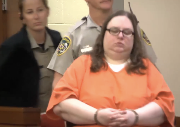 A detainee in orange attire is brought into a courtroom by an officer, suggesting the start of a legal proceeding.