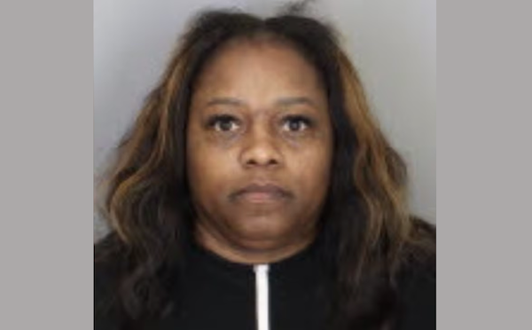 Image of a woman with loose, dark hair and a somber demeanor, positioned in front of a light-colored wall.