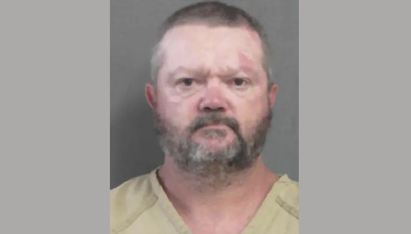 Head-and-shoulders image of a man in beige correctional attire, looking straight at the camera with a serious expression, set against a plain background.