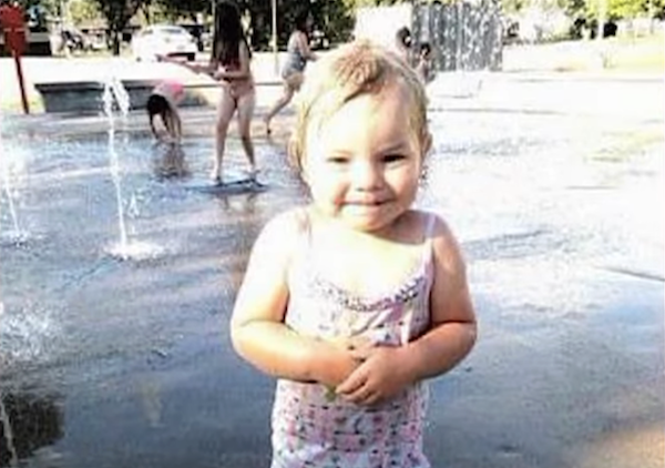 Smiling child wearing a patterned swimsuit at an outdoor splash park, with several kids playing and water features in the background.