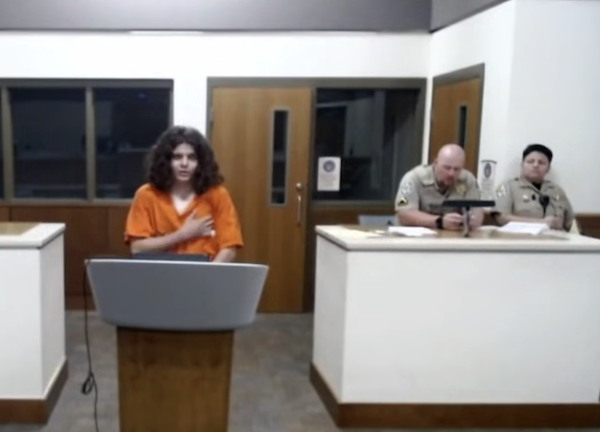 Person wearing an orange jail uniform stands alone at a lectern in a courtroom, while two sheriff’s deputies sit and observe from behind.