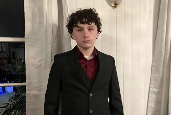 A curly-haired adolescent dressed in a suit stands against a bright white background, with a window showing night reflections and greenery beside him.