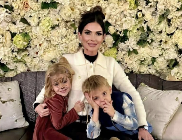 Three people sit closely on a wooden bench: an adult woman in a pale jacket, a girl with blonde hair and a butterfly painted on her face, and a boy in blue attire, all posing for a cheerful, cozy photo with a floral background.