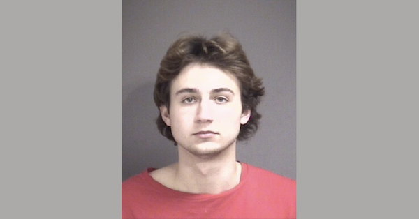 Arrest photo showing a man with medium-length hair and a serious demeanor, wearing a red shirt, set against a simple gray background.