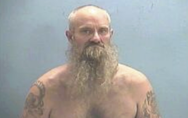 Bearded individual with prominent arm tattoos and a stern expression, captured in a mugshot with a solid light blue background.