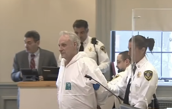 Inside a courtroom, a man in a white suit with hands cuffed behind his back is guided by a female officer, with other court staff and officers standing nearby.
