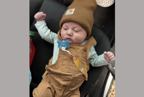 Baby in brown bib overalls and a tan beanie, with a pacifier on a strap, relaxes in a car seat surrounded by everyday items.