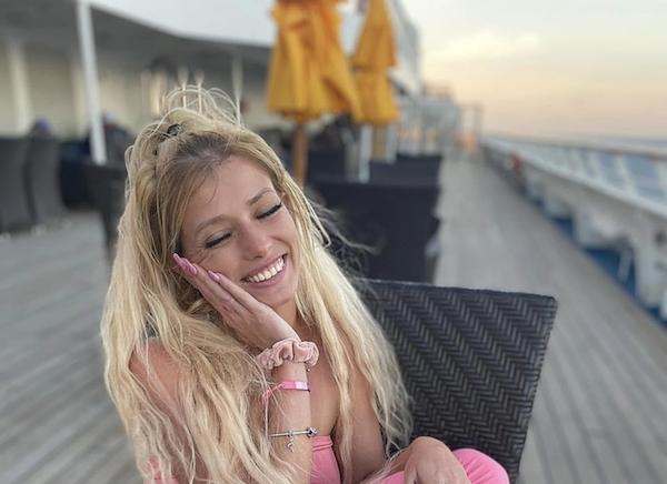 Cheerful young woman lounges on a deck chair aboard a ship, wearing a pink outfit and a scrunchie, surrounded by a wooden deck and leisurely atmosphere.