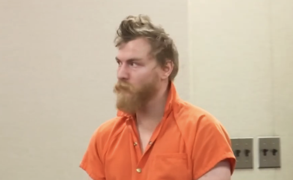 Bearded man in an orange inmate suit stands against a plain wall with two electrical outlets visible in the background.