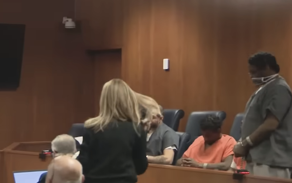 Three individuals in prison uniforms are seen appearing before the judge in a courthouse, with legal staff interacting nearby and a wooden wall behind them.
