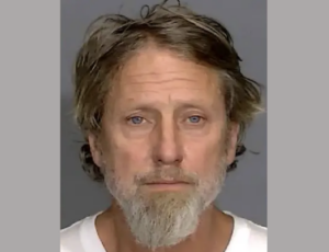 Head-and-shoulders image of a man with fair skin and a salt-and-pepper beard, positioned in front of a gray wall, wearing a white top.