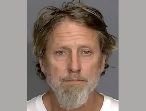 Head-and-shoulders image of a man with fair skin and a salt-and-pepper beard, positioned in front of a gray wall, wearing a white top.
