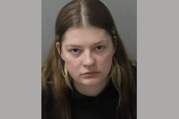 Portrait-style image of a woman with long brown hair and minimal makeup, looking straight into the lens, set against a simple background.