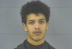 Portrait-oriented mugshot of a young man with dark curly hair and a neutral expression, photographed against a gray cinder block background.