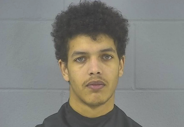 Portrait-oriented mugshot of a young man with dark curly hair and a neutral expression, photographed against a gray cinder block background.